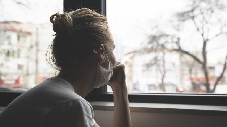 rear view closeup of young Caucasian woman wearing medical protection face mask watching the city by the window quarantine concept