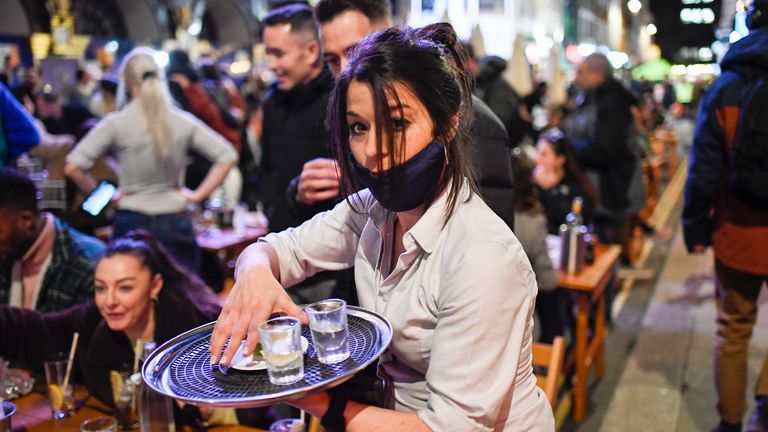 A waitress serves drinks at setup tables outside pubs in Soho, in London, on the day some of England's third coronavirus lockdown restrictions were eased by the British government, Monday, April 12, 2021. People across England flocked to shed shaggy locks and browse for clothes, books and other 