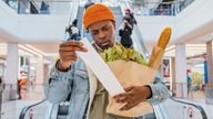 Surprised African-American man in denim jacket looks at receipt total in sales check holding paper bag with products in mall