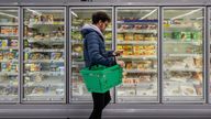 Man shopping in a supermarket while on a budget. He is looking for low prices due to inflation, standing looking at his phone in front of a row of freezers. He is living in the North East of England.

