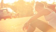 Exhausted woman sitting beside the road. She looks like she has been exercising or running and is sitting down for a rest. She is wearing sports clothing . Light is sunset or sunrise with a car in the back ground. Back lit with lens flare and copy space.