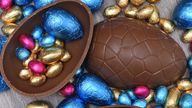 Pile or group of multi colored and different sizes of colourful foil wrapped chocolate easter eggs in pink, blue, and gold. Large halves of a brown milk chocolate egg have mini eggs inside, on a grey wooden background. 