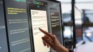 Close-up stock photograph showing a touchscreen monitor being used in an open plan office. A woman’s hand is asking an AI chatbot pre-typed questions & the Artificial Intelligence website is answering.