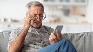 Poor Eyesight. Senior Man Squinting Eyes Reading Message On Phone Wearing Eyeglasses Having Problems With Vision Sitting On Couch At Home. Ophtalmic Issue, Bad Sight In Older Age Concept